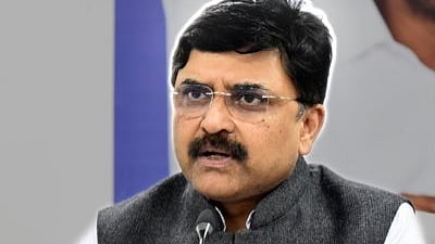 PV Ramesh is seated at a table and speaking through a mic. He is wearing glasses, a dark grey sleeveless jacket and a white shirt.