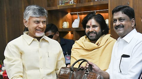 The image shows a group of men in a formal setting. One of them, AP CM Chandrababu Naidu wearing a cream-colored shirt, is smiling while receiving a brown leather briefcase from another man in a white shirt, state Finance Minister Payyavula Keshav. A third man, Deputy CM Pawan Kalyan, dressed in saffron robes with a red tilak on his forehead, is also present, smiling. The background features a wooden shelf with decorative items.