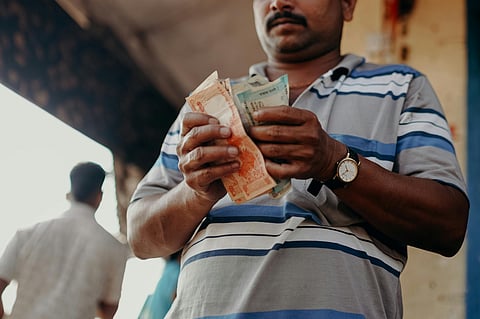A man wearing a striped polo shirt and a wristwatch is counting Indian currency notes. The focus is on his hands and the money, while his face is partially visible. The background is slightly blurred, showing other people and an outdoor setting.