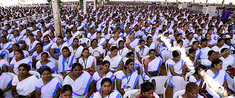 The image shows a large gathering of women wearing white sarees with blue borders, seated in an outdoor setting. Many of them have ID cards around their necks, suggesting they might be part of an organized group or workforce. The crowd appears attentive, possibly attending a conference, seminar, or a government-related event. The women in the image are ASHA (Accredited Social Health Activist) workers. 