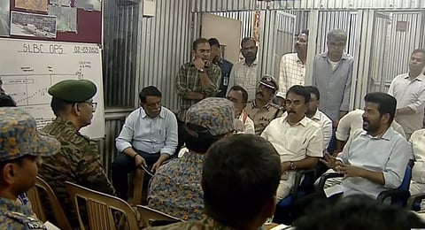 The image shows a meeting taking place inside a makeshift or temporary structure with corrugated metal walls. Several individuals, including government officials, police officers, and military personnel, are seated and engaged in discussion. A whiteboard in the background has "SLBC OPS" written on it, along with a date, "02-03-2025." The person speaking is Telangana Chief Minister Revanth Reddy, addressing the group while gesturing with his hands. The atmosphere suggests an emergency or strategic operations meeting, possibly related to a significant event or crisis.