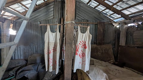 Inside of a tin shed. A wooden pole stands in the middle. On it, a stick has been tied horizontally and two white, sleeveless petticoats hang from either sides. There are hands painted in red on the white fabric. The shed is in a dilapidated state,. 