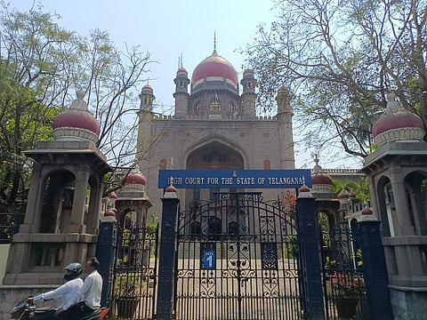 The image shows the entrance of the High Court for the State of Telangana in Hyderabad, India. The building features Indo-Saracenic architecture with red domes and intricate detailing. A blue sign with white lettering marks the court's name above the main gate, which is made of black wrought iron. The entrance is flanked by ornamental pillars topped with small domes. Trees partially frame the building, and two people on a motorcycle are seen in the foreground, adding a sense of movement.