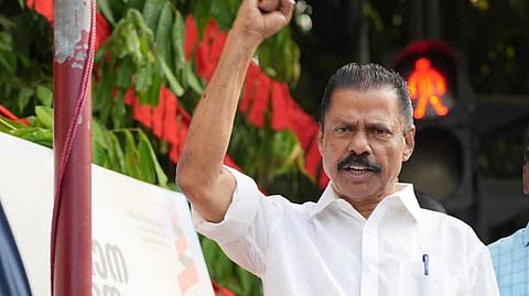 MV Govindan in a white shirt raises his fist in the air while speaking at a public event. Red flags are visible in the background.