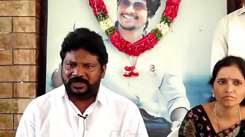 The image shows Pranay's father Balaswamy and mother Hemalatha seated in front of a large photograph of Pranay wearing sunglasses and smiling. The photograph is adorned with a garland. Balaswamy appears to be speaking emotionally, while Hemalatha looks solemn. The background suggests an indoor setting with a mix of brick and painted walls.
