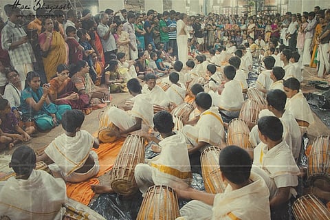 A group of children in traditional attire play drums in front of a gathered crowd at an indoor event.

