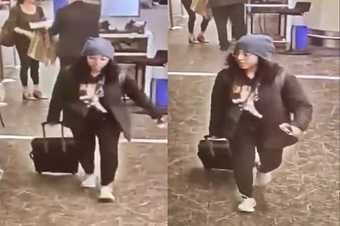 A woman dressed in an all black attire and a blue beanie, walking with a black suitcase.