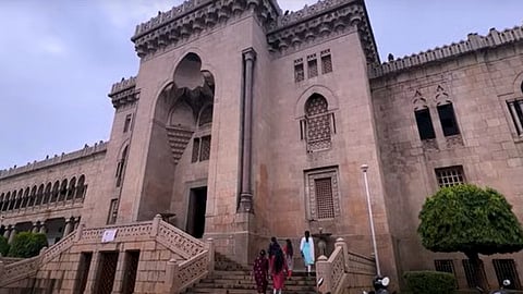 The image shows the historic Osmania University stone building with Indo-Saracenic architectural elements, featuring arched windows, intricate jali (lattice) work, and decorative parapets. A grand staircase leads up to the entrance, where a group of people dressed in traditional Indian attire is walking up. The sky is overcast, and the surrounding area is adorned with greenery.