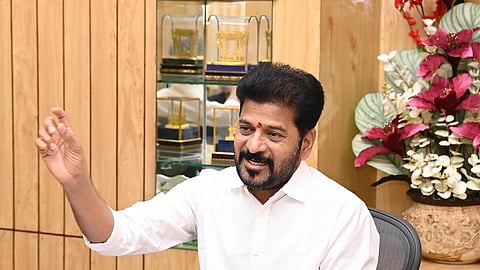 Telangana CM Revanth Reddy, wearing a white shirt, is sitting at a desk in an office. He is gesturing with his right hand while speaking and has a friendly expression. Behind him, there is a wooden-paneled wall, a glass display case with awards or trophies, and a floral arrangement with red and white flowers. There are office supplies like a notebook and pen on the desk.