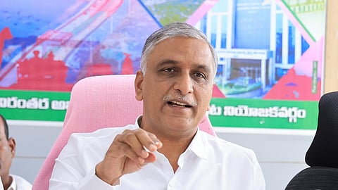BRS leader T Harish Rao is seen speaking at a press conference, dressed in a white shirt. He is seated in a chair with a pink cover, gesturing with his right hand while addressing the media. Multiple microphones with various news channel logos are placed in front of him. Behind him, a colorful banner with Telugu text and images of infrastructure projects is visible. The setting appears to be an official or government office.