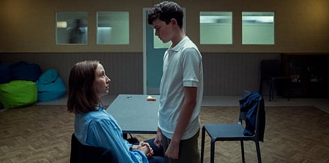 A tense, emotional scene in a dimly lit room where a seated woman in a light blue shirt looks up at a teenage boy standing in front of her. The boy, dressed in a white polo and green trousers, holds her hand as they exchange a serious, intense gaze. The room has neutral-colored walls, windows at the top, and scattered bean bags and chairs, suggesting a clinical or institutional setting.