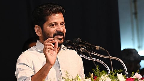 The image shows Telangana CM Revanth Reddy in a light-colored shirt speaking at a public event. He is standing behind a podium decorated with colorful flowers and is gesturing with his hand while addressing an audience. There are multiple microphones in front of him, and the background is dark with some people visible.
