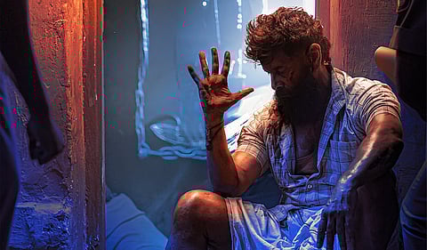 A rugged man with a thick beard and unkempt hair sits on the floor of a dimly lit room. His clothes are tattered and stained, and his muscular arms bear visible wounds. He raises his hand, seemingly in a moment of exhaustion or contemplation. The lighting is dramatic, with warm orange tones on one side and cool blue hues on the other, adding to the intense and moody atmosphere. Chains are visible in the background, suggesting a confined or prison-like setting.