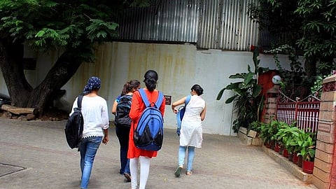 A group of five young women, dressed in casual and traditional Indian attire, walk together on a paved pathway. They carry backpacks and shoulder bags. The background features a large tree, a metal-roofed building, and a gated entrance adorned with potted plants. The setting appears to be an educational institution or a similar public space.