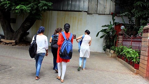 A group of five young women, dressed in casual and traditional Indian attire, walk together on a paved pathway. They carry backpacks and shoulder bags. The background features a large tree, a metal-roofed building, and a gated entrance adorned with potted plants. The setting appears to be an educational institution or a similar public space.