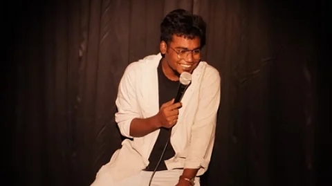 Manjeet Sarkar at a comedy gig, seated on a stool, wearing white pants and shirt and a black tee inside the shirt. He is holding a wired mic in his right hand and his left hand is resting on his left knee. The stage has a black backdrop.