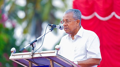 The image shows Kerala Chief Minister Pinarayi Vijayan, wearing a white shirt and glasses, speaking into a microphone at an outdoor event. He is standing behind a metal podium with a blue sign attached to it. The background features red fabric drapery and blurred greenery.