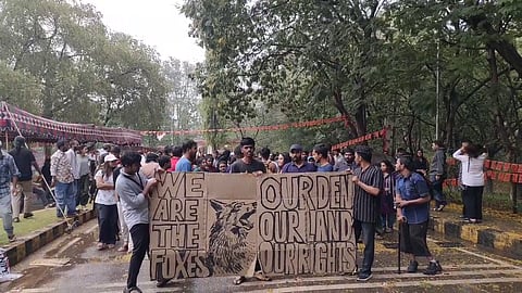 A group of people is gathered outdoors in a protest. Several individuals are holding a large cardboard sign with bold, hand-drawn lettering and an illustration of a fox. The sign reads:

"WE ARE THE FOXES"
"OUR DEN, OUR LAND, OUR RIGHTS"

The setting appears to be a university or public park, with trees, tents, and banners in the background. Many protesters are engaged in conversations, and some are looking at the sign. The atmosphere suggests activism, possibly related to land or student rights.