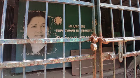 A poster of late Tamil Nadu Chief Minister J Jayalalithaa that reads "Amma Kudineer" with machinery for providing water next to it, locked up behind a rusty gate.