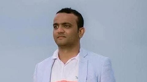 Vinay Somaiah with short hair, wearing a pastel-colored polo shirt and a light blue blazer, stands outdoors against a blurred background of the sky and possibly a beach. He has a neutral expression and is gazing into the distance.