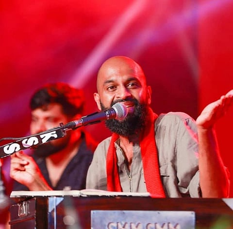 Singer Aloshi Adams performing on stage, wearing a grey kurta and a red scarf. He is singing into a microphone with an expressive gesture, raising one hand. The stage is bathed in red lighting, and a musician is visible in the background.