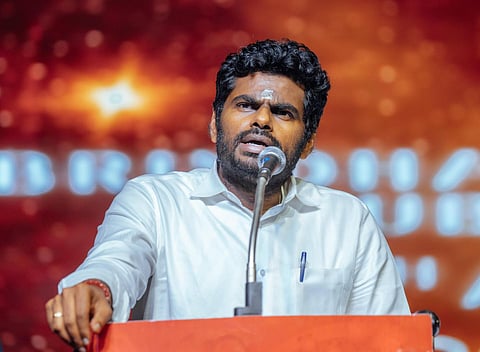 K Annamalai, the Tamil Nadu state BJP president, speaks into a microphone at a public event. He is wearing a white shirt with a pen clipped to his pocket and has a serious, focused expression. The background features a blurred, warm-toned stage with illuminated text.