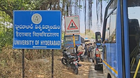 A signboard at the entrance of the University of Hyderabad displays the university's name in Telugu, Hindi, and English. Nearby, a "Pedestrian Crossing" sign is also visible. The road beside the sign is crowded with blue buses, cars, and motorcycles, with people seen walking among the vehicles. Large tree roots hang overhead, and the surroundings are dry with sparse vegetation.