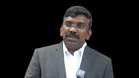 Reny Wilfred wearing a dark grey suit and white shirt, with a microphone clipped to his lapel, standing against a plain white background.
