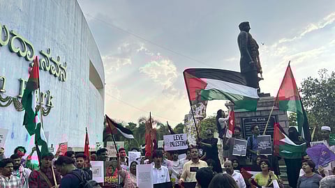 A large group of protesters holding Palestinian flags and placards gather near a statue at Freedom Park in Bengaluru. Some people are standing on the statue’s base, waving flags and chanting slogans. Placards read messages like “Student Intifada,” “Leve Palestina,” and “Education is learning about the oppressor’s history.” The backdrop includes a curved wall with Kannada and English signage, palm trees, and a partly cloudy sky.