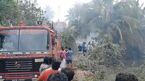 A red fire truck is seen on a narrow rural path surrounded by fallen tree branches and dense smoke. Several people are gathered around, with some wearing casual clothes, appearing to observe or assist in a rescue or emergency response. Palm trees and vegetation are visible in the background, suggesting a tropical setting. The atmosphere is hazy, indicating a recent fire or explosion in the area.