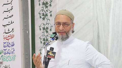 AIMIM president Asaduddin Owaisi, wearing a white kurta, light olive cap, and glasses, is speaking at a podium with a microphone. The podium has a "4tv News" mic attached. He has a grey beard and is gesturing with his hand while speaking. Behind him is a wall with decorative green vine patterns and text written in Urdu.