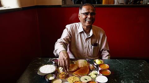 A man is seated smiling and in front of him on a granite table is the thaali at MTR with various dishes arranged neatly on a steel plate