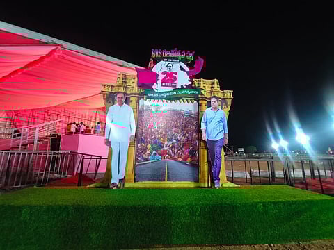 A brightly lit stage setup at night with pink drapes and a green turf base, featuring large cutouts of two men, KCR in a white outfit and KTR in a blue shirt and dark pants. They stand on either side of a decorated frame showcasing a massive crowd scene and a banner reading 'BRS 25 Years' in Telugu, celebrating the party's silver jubilee.