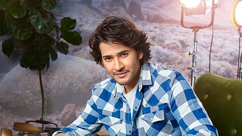 Mahesh Babu with tousled dark hair and a slight smile sits casually on a green armchair. He’s wearing a blue and white checkered shirt over a white T-shirt paired with blue jeans. Behind him, there are bright studio lights, a leafy plant, and a dreamy, misty backdrop with rocky formations.