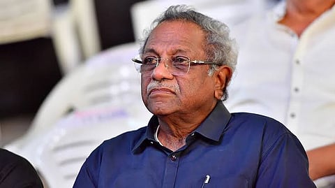 An elderly man with gray curly hair and glasses, dressed in a dark blue shirt, sitting on a white plastic chair, looking thoughtful at an event.