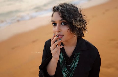 Prayaga Martin standing on a sandy beach, wearing a black outfit with layered green necklaces, looking thoughtfully at the camera with her hand near her lips, soft waves visible in the blurred background

