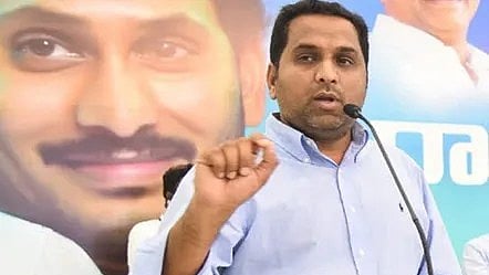 A man in a light blue shirt is speaking into a microphone at a podium. Behind him is a large poster featuring a close-up of YS Jagan's face smiling. The background suggests a political event or press conference.