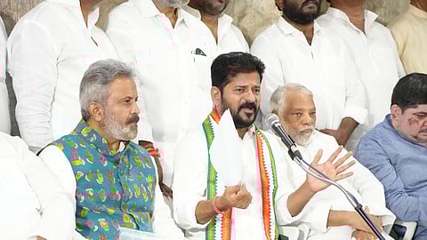 A group of Indian political leaders is seen seated during a press conference. The central figure, Telangana CM Revanth Reddy, wearing a white shirt and a tricolor party scarf, is speaking into a microphone and holding papers, gesturing with his hands. To his left, another leader is wearing a colorful Nehru jacket with hand symbols. Most other attendees are dressed in white, indicating a formal or political setting.