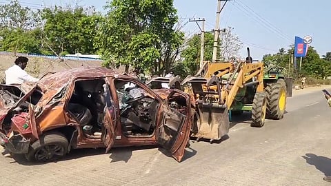 A severely damaged car with its doors open is being lifted by a tractor on a sunny day. The car appears completely crushed, indicating a serious accident. Several people are around the vehicle, including a man operating the machinery and another photographing the scene. Trees, electrical poles, and a fuel station sign are visible in the background.