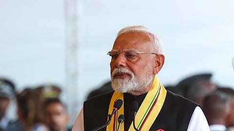 Indian Prime Minister Narendra Modi stands at a podium decorated with traditional Indian motifs and floral arrangements, delivering a speech. Yellow marigold flower garlands hang above him, and he wears a formal black and white outfit with a yellow ceremonial sash and red rosette.