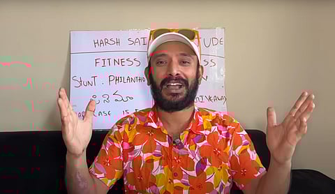 A man wearing a colorful floral shirt, white cap, and orange sunglasses (resting on the cap) is sitting on a black couch with his hands raised mid-gesture. Behind him is a white poster board with handwritten text in English and Telugu, including words like “HARSH SAI,” “FITNESS,” and “PHILANTHROPY.” The setting appears casual, likely part of a YouTube video or vlog.