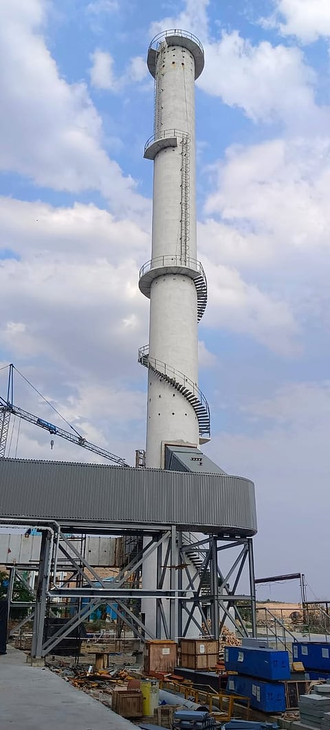 A tall industrial chimney structure at a construction site, likely part of a waste-to-energy or similar facility. The chimney is cylindrical, made of concrete, and features a spiraling metal staircase wrapping around its exterior. It rises from a metal platform structure with support beams and is surrounded by construction equipment, crates, and scattered materials. A crane and partly cloudy sky are visible in the background.