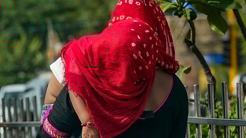 A woman wearing a red dupatta and black traditional attire carries a child in her arms on a sunny day, walking along a metal fence lined with greenery. The child is dressed in white and appears to be resting on her shoulder.