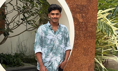 dil standing in front of a circular architectural feature on a reddish-brown wall with tropical plants in the background. He is wearing a green and white palm-print shirt with the sleeves rolled up and black pants, posing with one hand in his pocket and a slight smile.
