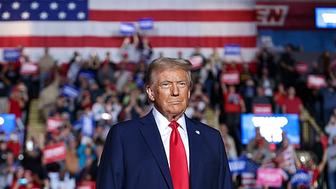 Donald Trump stands in front of a large crowd at a political rally, with a giant American flag hanging behind him. He is wearing a navy blue suit, white shirt, and red tie, looking forward with a serious expression. The crowd in the background holds up signs in red, white, blue, and pink.