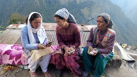 Three women sit on a low wall with hills seen in the background. The woman on the extreme left is looking at the mobile phone in her hand as the woman in the middle also peeks into it.