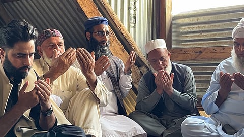 A group of men sit in solemn prayer inside a modest room with corrugated metal walls. Their hands are raised in Islamic supplication. The man wearing a blue cap and glasses, seated in the center, is Iqbal’s brother, Farooq.