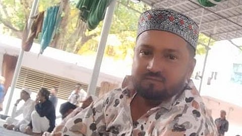 A man wearing a patterned cap and a multicolored, abstract-print shirt is seated indoors on a mat among a group of people. The setting appears to be a community or religious gathering, with natural light filtering through the open-sided structure and green cloth decorations hanging from the ceiling.