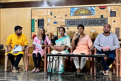 Five people sit at a table on a stage with a "PRESS CLUB OF BANGALORE" backdrop. Individuals in a yellow kurta, pink top, sari, peach top speaking into a microphone, and a grey shirt are visible.
