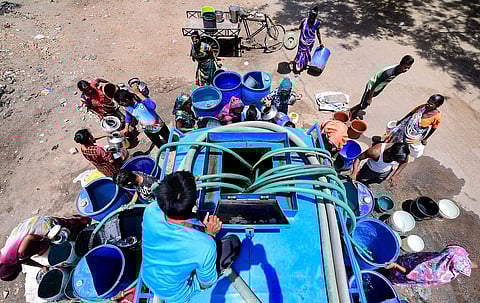 A group of people gather around a blue water tanker on a hot day to collect water in various containers, including plastic buckets and metal pots. The photo, taken from above, shows hoses from the tanker filling the vessels as people wait and manage their positions. The scene reflects a water scarcity situation, with the setting appearing to be a dry, urban or semi-urban area.